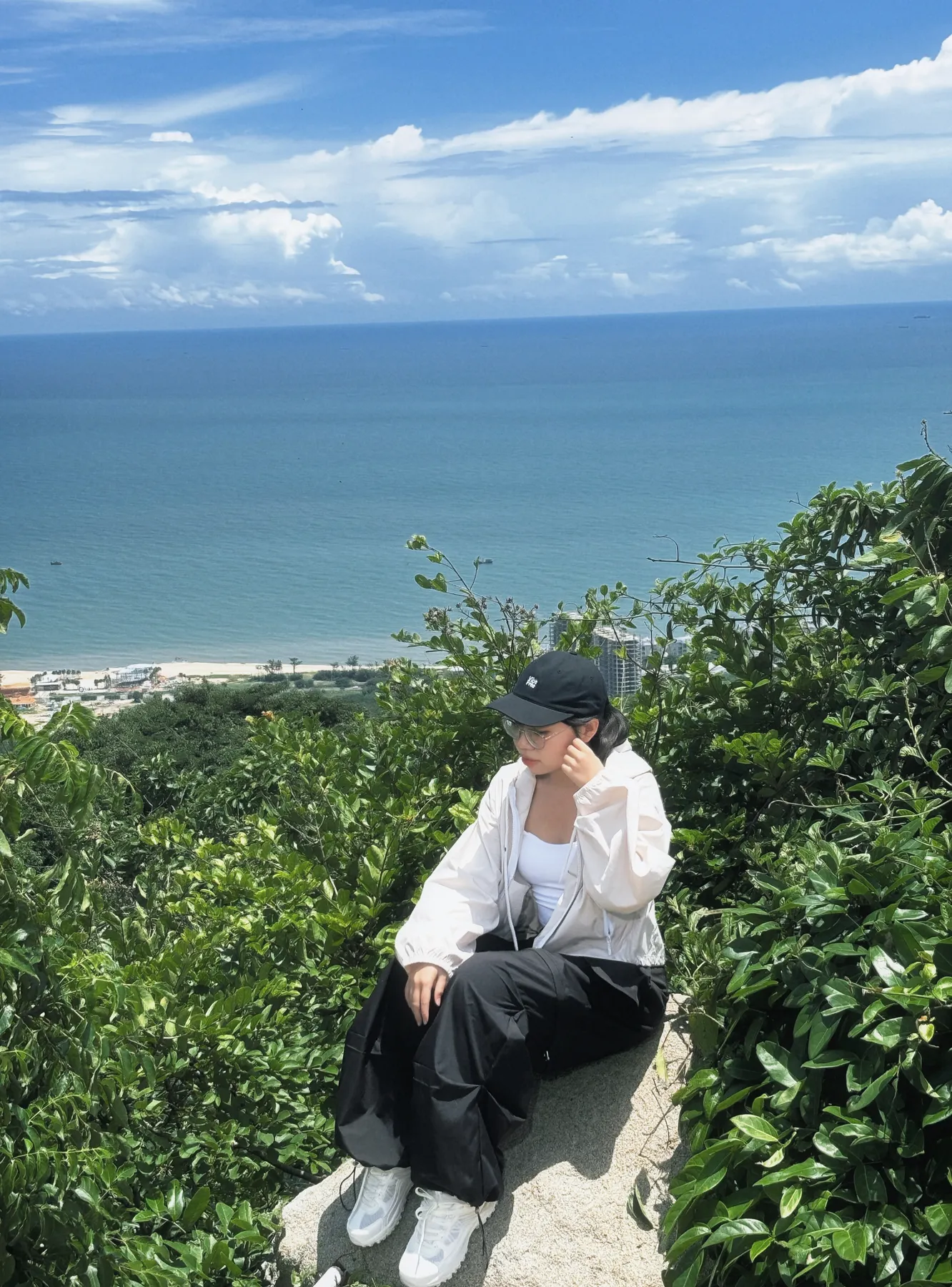Người mặc quần đen và áo khoác sáng ngồi trên đá, phía sau là cây xanh và biển.