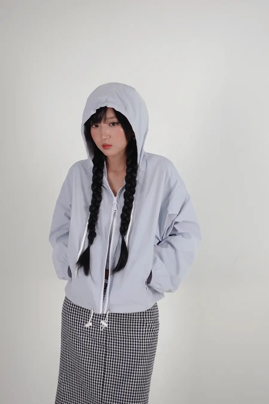 Áo khoác dù màu xanh nhạt có mũ trùm, phối chân váy kẻ caro, chụp trong studio