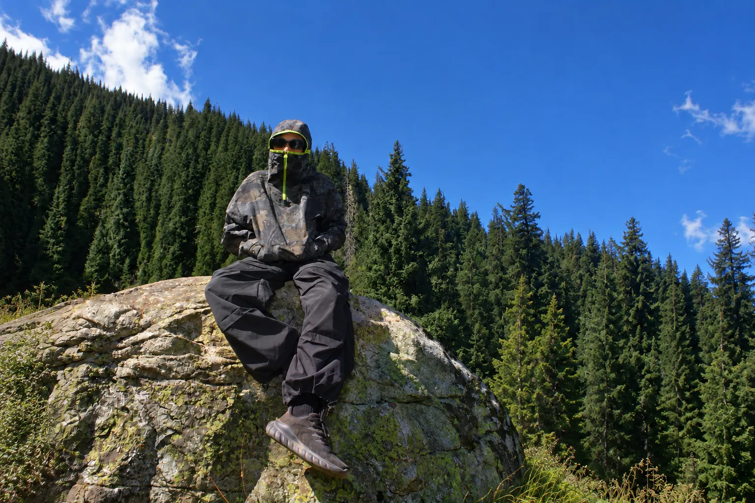 Người đi trekking đội mũ trùm, mặc quần cargo ống rộng màu đen, ngồi trên tảng đá giữa rừng thông dưới trời xanh (ảnh mở rộng bằng AI từ ảnh thật)